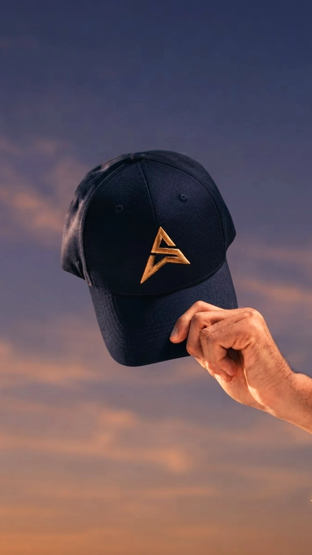 A navy cap with the gold embroidered Awesome A mark held up against a dusk sky